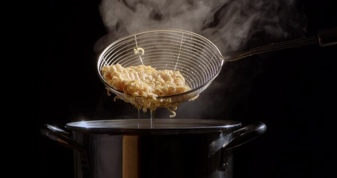 Slow motion scene while the cook is using a strainer to boil yellow noodles in a stainless steel pot that is boiling and steaming.