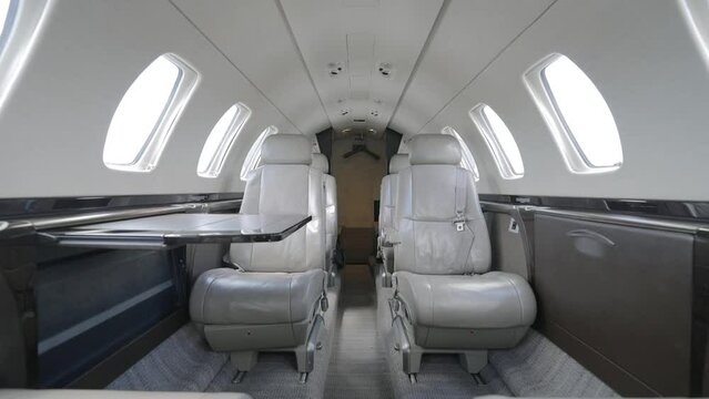 Contemporary interior of private jet cabin. Four white leather seats with headrests arranged in pairs on either side of narrow aisle. Light grey carpet and walls