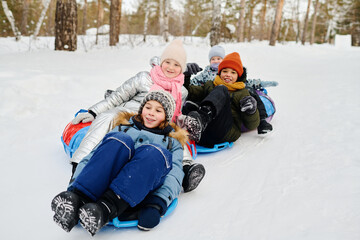 Group of cute laughing children in winterwear sitting on slides and tubes while moving down snow hill at winter resort or forest