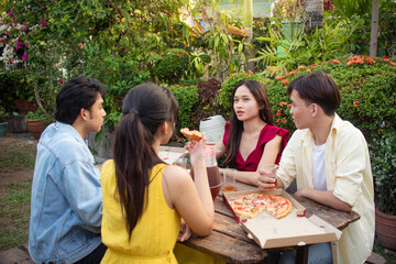 Four young asian friends enjoying pizza and drinks during an outdoor picnic in a lush garden or lawn. Officemates catching up after a long time.