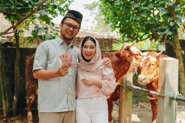 Cheerful young Asian Muslim couple holding money to buy cows cattle for sacrifices. Eid Al Adha...