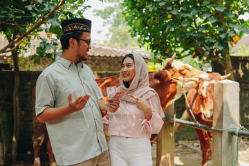 Excited young Asian Muslim couple pointing the money transaction they hold for cows cattle...
