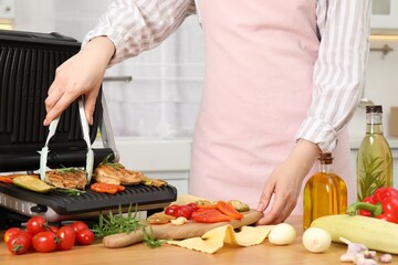 Woman cooking different products with electric grill at wooden table in kitchen, closeup