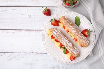 Delicious eclairs filled with cream, strawberries and mint on white wooden table, top view. Space...