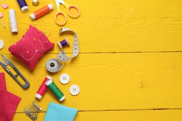 Red pincushion with pins and other sewing tools on yellow wooden table, flat lay. Space for text