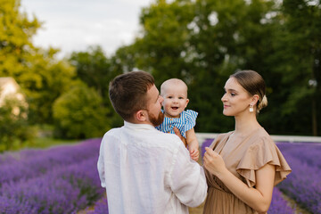 Fototapeta premium Family with a baby on a lavender field. A woman in brown dress and a man in white skirt with a beard are holding a newborn baby girl in a blue dress in a lavender field. Family, lifestyle concept.