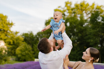 Fototapeta premium Father is lifting up his daughter in the air while they have fun together in the lavender field. Man and woman playing with his newborn baby girl in blue dress on the lavender field.