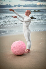 Full length portrait of hairless girl with alopecia in white futuristic suit standing on sea beach...