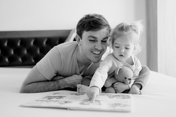 Little girl runs her finger along the lines of the book that her dad reads to her, lying on the...