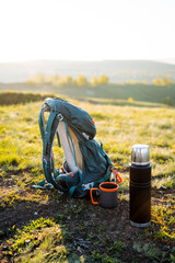 Outdoor backpack and thermos set in scenic landscape for adventurous hiking trips. Enjoy a cup of refreshment in natures beauty grassy fields, mountains, and sunset views