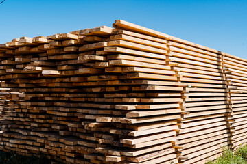 A stack of freshly cut wooden planks sits under a clear blue sky at an outdoor sawmill, showcasing...