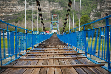 A suspension footbridge with blue railings and wooden planks offers a scenic pathway for...