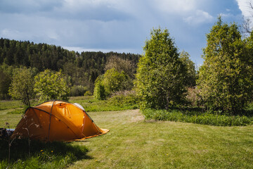 Amidst a lush green forest meadow stands a bright orange tent under a blue sky, creating a serene...