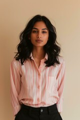 Obraz premium South Asian woman with wavy, shoulder-length black hair, wearing a pink and white striped button-up shirt and black trousers, beige background