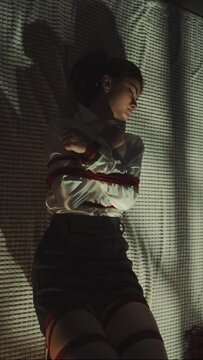 Vertical screen: Zoom out woman bound with red shibari ropes, lying on white blanket in softly lit room, traditional Japanese rope, aesthetics and calm. White sneakers nearby, intimate scene