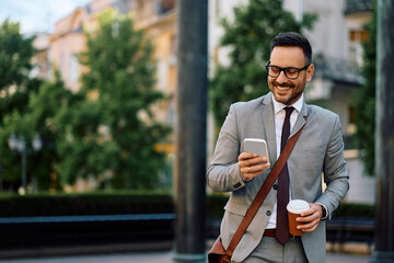 Happy businessman text messaging on cell phone while drinking coffee in city.