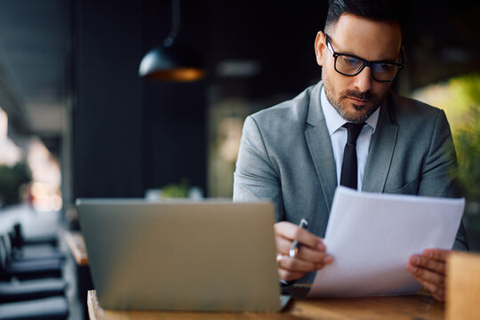 Male entrepreneur going through business reports while working on laptop in caf&eacute;.