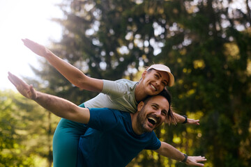 Cheerful athletic couple piggybacking with arms outstretched in park.