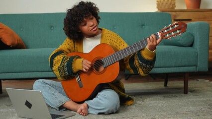 African American woman learning guitar watching tutorial on laptop at home. Black girl playing guitar singing song learning online music lessons. Artistic woman playing acoustic guitar learning chords