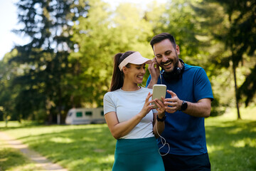 Happy couple of athletes listening music on earphones while using cell phone during outdoor workout.