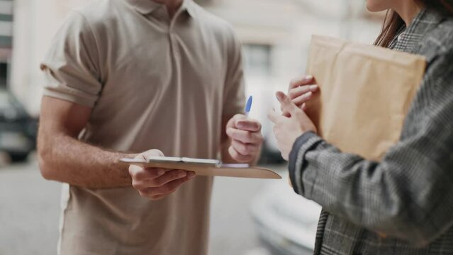 A cropped view of a courier man delivery of parcel while a customer signs for delivery. The woman, standing outdoors indicating a successful delivery process