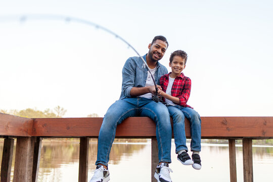 African American man and child sitting on a wooden pier holding fishing rods on the river, dad teaching his son to fish in the lake, family resting and relaxing on the weekend