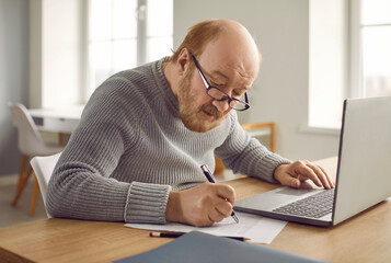Focused senior man making notes on paper and working with laptop. Portrait of elderly man in...