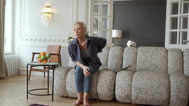 Woman in 60s sitting on couch in living room with laptop. She works or enjoys leisure online activities. tries to stand up and feels unexpected sharp back pain causes her to frown and hold her back