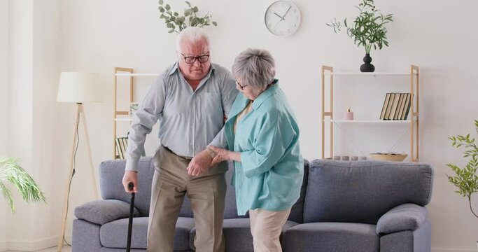 Married senior couple overcoming health problems together. Old woman helps sick weak man to stand up from sofa and walk slowly with crutch in living room. Disability, care, assistance, support concept