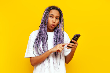surprised african american woman with colored dreadlocks using smartphone in shock on yellow...