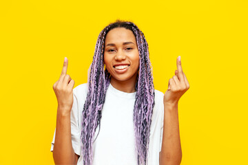cheerful african american woman with colored dreadlocks showing middle finger on yellow isolated...