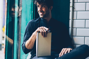 Smiling young man dressed in black shirt looking out of window holding notepad with mock up area...