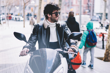 Cheerful hipster guy in black sunglasses parking own motorcycle on pavement in urban...