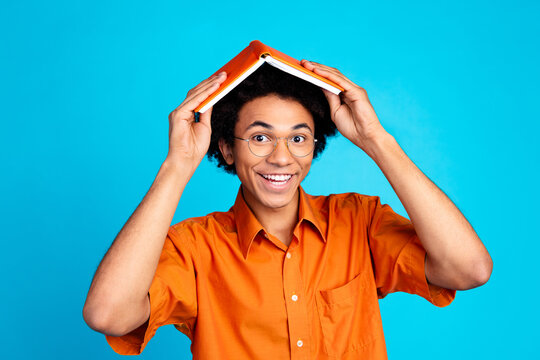 Photo of cheerful positive guy dressed orange shirt holding head book empty space isolated blue color background