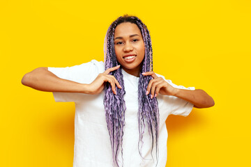 young african american woman with dreadlocks holding her hair in her hands on a yellow isolated background, girl with a unique hairstyle and colored braids smiling