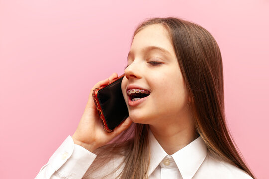 cheerful little teenage girl with braces talking on the phone on a pink isolated background, child communicating via mobile phone