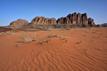 Ein Panorama des markanten Al Qattar Massiv in der Wüste des Wadi Rum in Jordanien bei strahlen blauem Himmel und im Vordergrund sind die typischen Sanddünen.