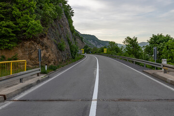 captures a serene, overcast day on a mountain road. The asphalt road curves gently to the right, bordered by a yellow and white guardrail on the left and a series of safety barriers on the right.