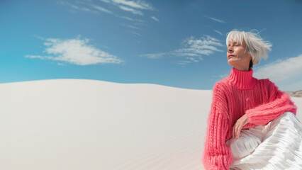 Fabulous at any age concept. Portrait of fashionable female mature model with short silver hair sitting in luxurious clothing among sands under the blue sky. Luxury brands campaign style