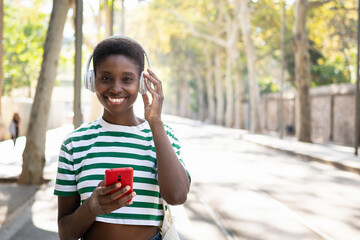 Young African woman listening to music with headphones and wireless red cell pone at city park 