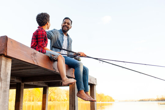 african american father and son barefoot fishing in the lake and sitting on a wooden pier and holding a fishing rod outdoors in summer, a man teaches a child to fish and relax outdoors on a river