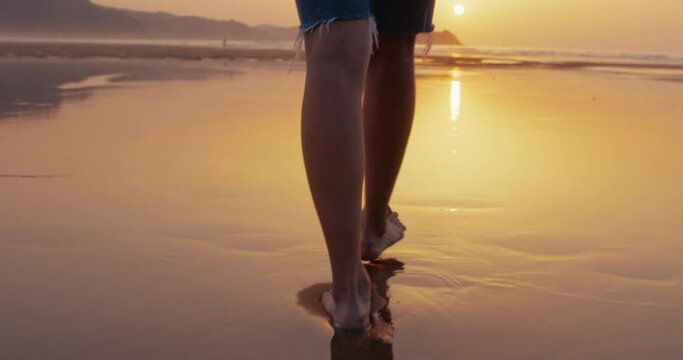 Close up on female feet step on low water reflecting setting sun on an amazing beach with mountains in background. Young woman on solo vacation travel. Moody and atmospheric self care concept