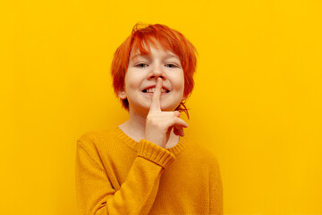 red-haired boy child shows a finger near his lips and a gesture of secrecy on a yellow isolated...