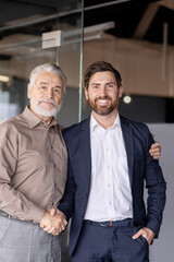 Senior businessman and young colleague happily shaking hands in a modern office, showcasing teamwork and collaboration