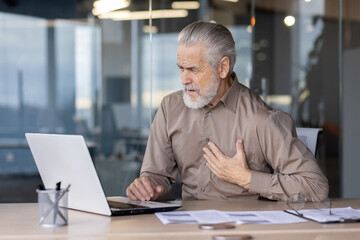 Older man, grey-haired, distressed, holding chest, experiencing pain while working on laptop in modern office setting with glass partitions and paperwork. Concept of health issues at work.