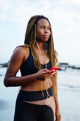 Sportive black female jogger dressed in active clothing listening music in earphones connected to phone strolling on seacoast in morning.Afro american woman with dreads wearing tracksuit for running
