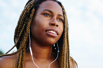 Afro american young woman with dreads listening audio songs from own player downloaded on smartphone walking outdoor on coasline.Dark skinned jogger enjoying music in earphones connected to cellular