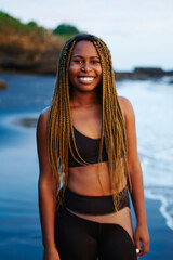 Portrait of positive afro american sportwoman with dreads standing on shore washed by ocean...