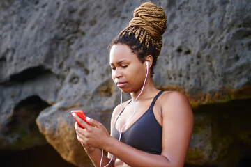 Afro american sportive girl choosing music application on smartphone for morning training on seashore.Young woman in active wear listening audio songs in earphones connected to cellular during workout