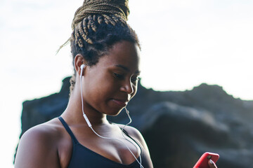 Afro american sportive woman choosing audio songs in music application on smartphone for morning run along seashore.Dark skinned female in tracksuit listening player downloaded on telephone device